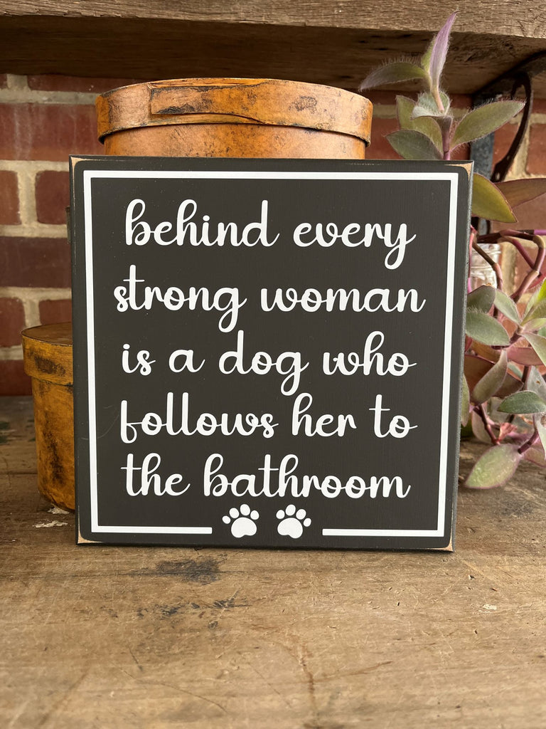 Square worn finish 8x8 inch black wood sign. Crisp white letters saying Behind every strong woman is a dog who follows her to the bathroom.  Two small paw prints at the bottom of the sign.