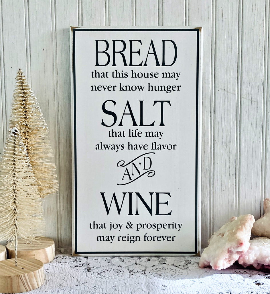 A rectangular white worn finishwood sign with the home blessing from the Christmas movie It's a Wonderful Life. Bread, that this house may never know hunger. Salt, that life may always have flavor. And Wine, that joy and prosperity may reign forever. Black lettering, Sign is available in several sizes. 